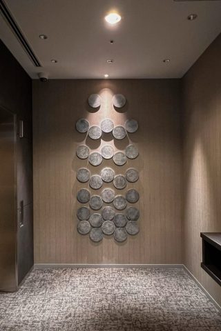 シルバーの特殊塗装を施した木製の円形パーツを情報に向かって立体的に配置した壁面アート。ホテルのエレベーターホールの壁面に設置された壁面装飾。